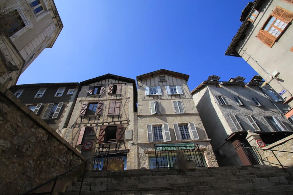Vieilles bâtisses traditionnelles avec volets divers, escalier en pierre et rampe, sous ciel bleu clair. Boutiques dont "Bio Diététique". Panneau sens interdit.