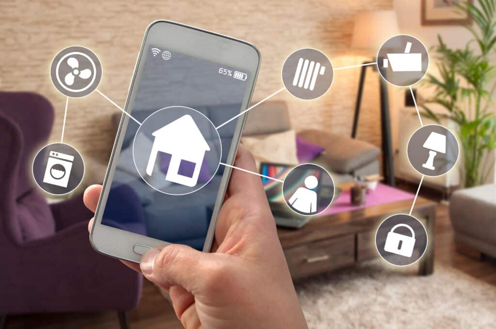 Main tient smartphone contrôlant domotique. Icônes ventilateur, lave-linge, chauffage, caméra, lumière, serrure. Batterie 65%. Intérieur maison flou.
