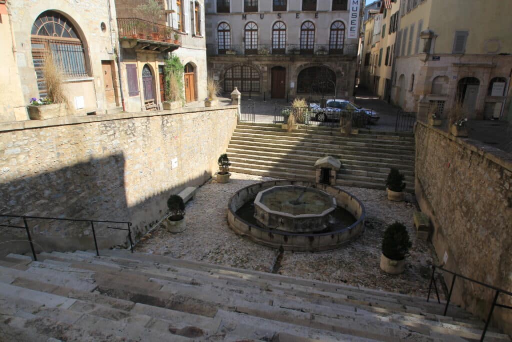 Place de village pavée avec fontaine octogonale, grand escalier et bâtiments anciens dont le MUSEE MUNICIPAL URBAIN CARRIOL.