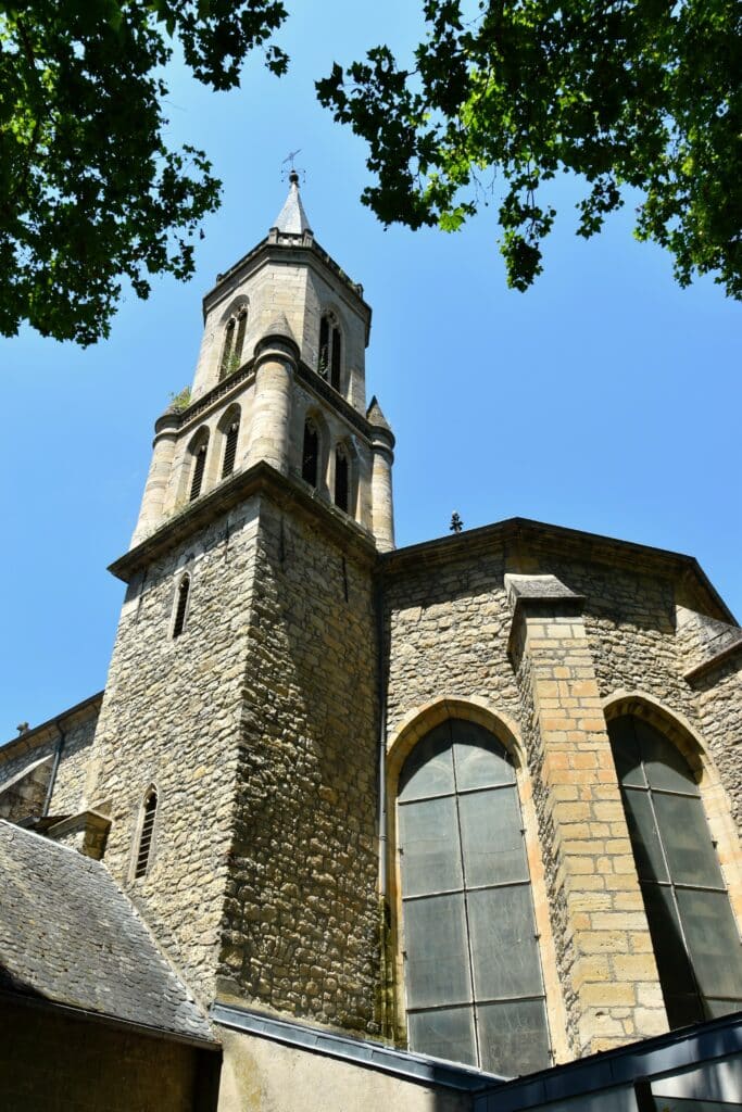 Vue en contre-plongée d'une tour d'église en pierre avec clocher, entourée de feuillage vert sur ciel bleu clair.